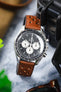Close-up of an Omega Speedmaster Professional chronograph watch featuring a black dial with three white chronograph sub-dials, black tachymeter bezel, and polished stainless steel case, fitted with a cognac brown perforated leather rally strap with matching brown stitching. Displayed beside a black travel case on a marble-effect surface, the image highlights the strap's vintage motorsport-inspired design, premium handcrafted leather construction, and rich warm-toned finish.