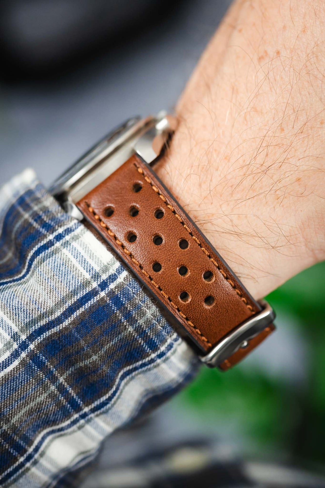 Close-up of a brown perforated leather watch strap worn on a wrist, showing detailed stitching and a silver buckle. The strap is paired with a watch partially out of frame, worn over a blue-and-white plaid shirt cuff, with soft-focus greenery in the background.