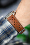 Close-up of a brown perforated leather watch strap worn on a wrist, showing detailed stitching and a silver buckle. The strap is paired with a watch partially out of frame, worn over a blue-and-white plaid shirt cuff, with soft-focus greenery in the background.