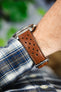 Close-up of a cognac brown perforated leather rally watch strap worn on the wrist, featuring matching brown stitching, multiple motorsport-inspired perforations, a polished stainless steel buckle, and a rich smooth leather finish. Paired with a blue plaid shirt cuff, the image highlights the strap's premium handcrafted construction, comfortable fit, and vintage racing-inspired design.