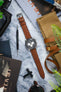 Flat lay of a chronograph wristwatch with a black dial and three white subdials on a brown perforated leather strap, arranged diagonally on a marble surface. Surrounding items include a tan canvas watch roll with leather straps, a spring bar tool labeled “WATCH OBSESSION,” sunglasses, printed magazines with visible “ORACLE” text, and green plant leaves partially framing the scene.