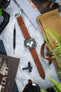 Omega Speedmaster Professional chronograph watch with a black dial, three white chronograph sub-dials, black tachymeter bezel, and a cognac brown perforated leather rally strap with matching brown stitching, displayed on a marble-effect surface alongside a Watch Obsession spring bar tool, canvas pouch, sunglasses, magazines, and greenery, highlighting the strap's vintage motorsport-inspired design and premium handcrafted leather construction.