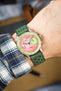 Close-up of a wrist wearing a mechanical chronograph watch with a vibrant pink textured dial and light green outer ring, featuring two subdials and black hands. The watch is paired with a green perforated leather strap and worn over a blue-and-white plaid shirt cuff, with a softly blurred indoor background.