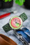 Close-up of a mechanical chronograph wristwatch with a vivid pink textured dial and light green outer ring, featuring two subdials and black hands. The watch is fitted to a green perforated leather strap with matching stitching and is propped against a metal multi-tool on a marble surface, with blurred greenery and watch accessories in the background.