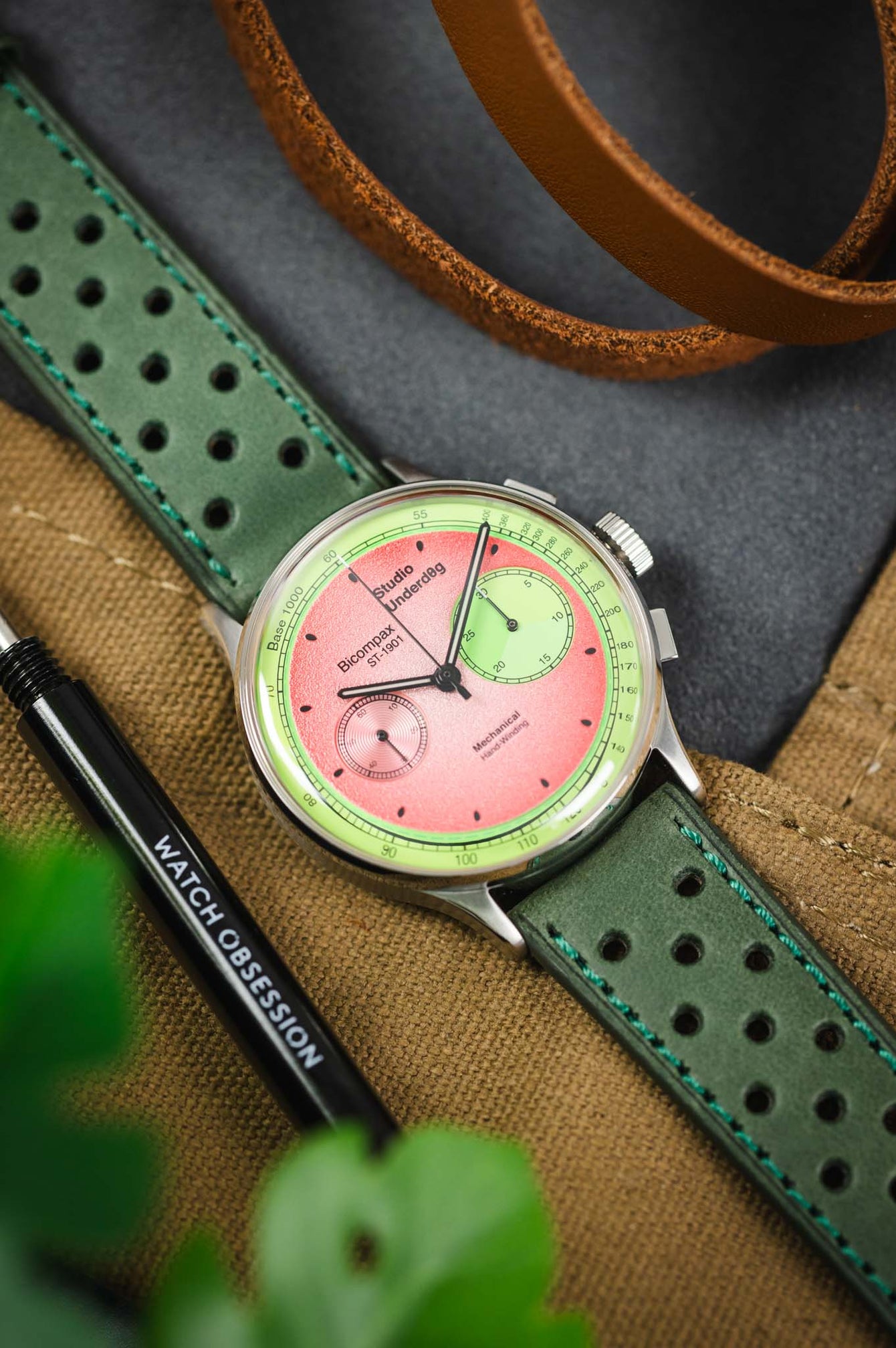 Close-up of a mechanical chronograph wristwatch with a vibrant two-tone dial in pink and green, featuring two subdials and black hands. The watch is mounted on a green perforated leather strap with contrasting stitching and rests on a tan canvas surface alongside a brown leather strap and a spring bar tool labeled “WATCH OBSESSION,” with soft-focus green leaves partially visible in the foreground.