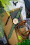 Flat lay of a mechanical chronograph wristwatch with a pink textured dial and light green outer ring, paired with a green perforated leather strap, placed diagonally on a marble surface. Surrounding items include a tan canvas watch roll with brown leather straps, a spring bar tool labeled “WATCH OBSESSION,” a black tool, camera equipment, and green plant leaves partially framing the scene.