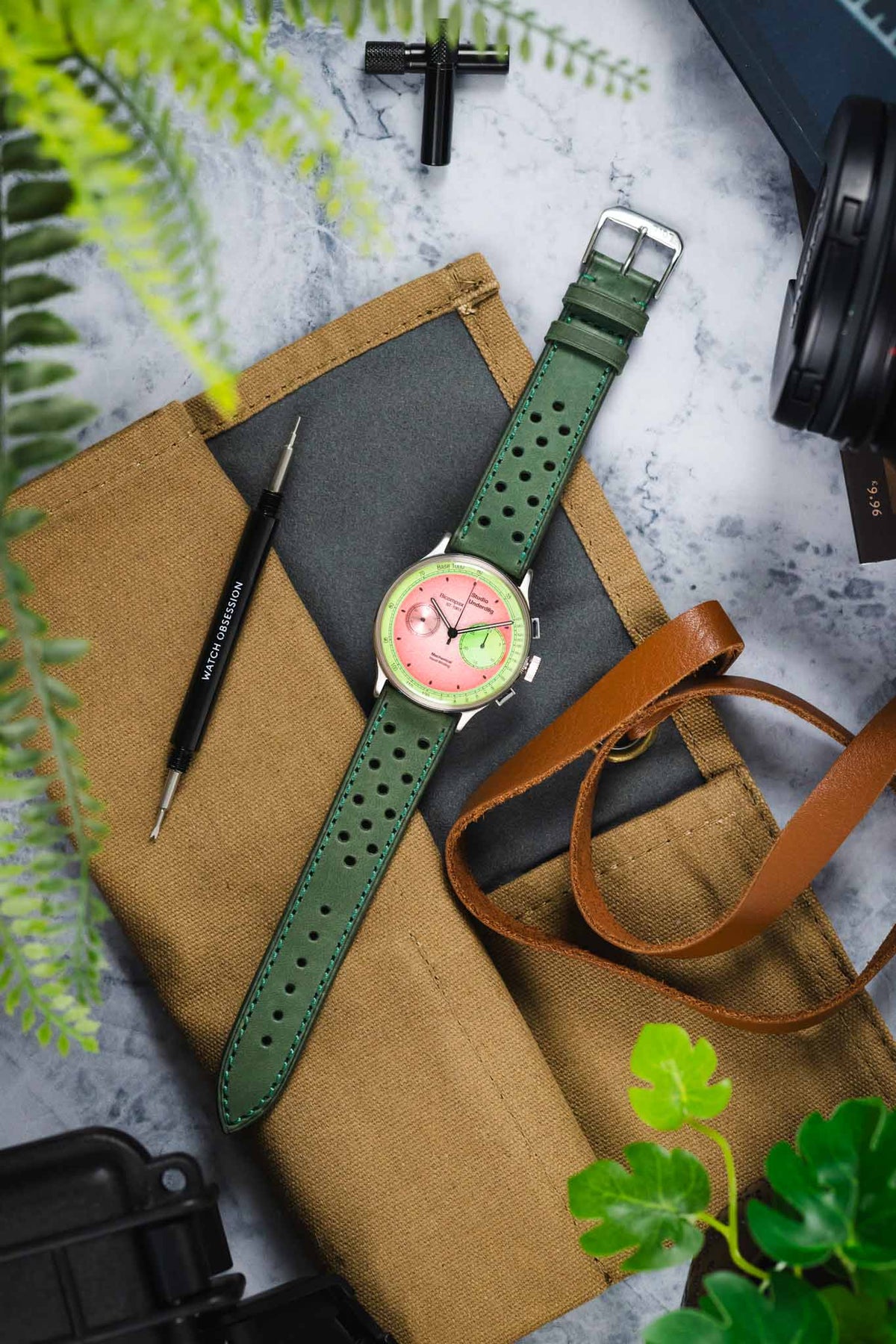 Studio Underd0g Bicompax chronograph watch with a pink dial and light green sub-dial, fitted with a forest green perforated leather rally strap featuring turquoise contrast stitching and a tapered profile, displayed on a canvas tool roll alongside a Watch Obsession spring bar tool, camera lens, leather accessories, and greenery, highlighting the strap's vintage motorsport-inspired design and premium handcrafted construction.