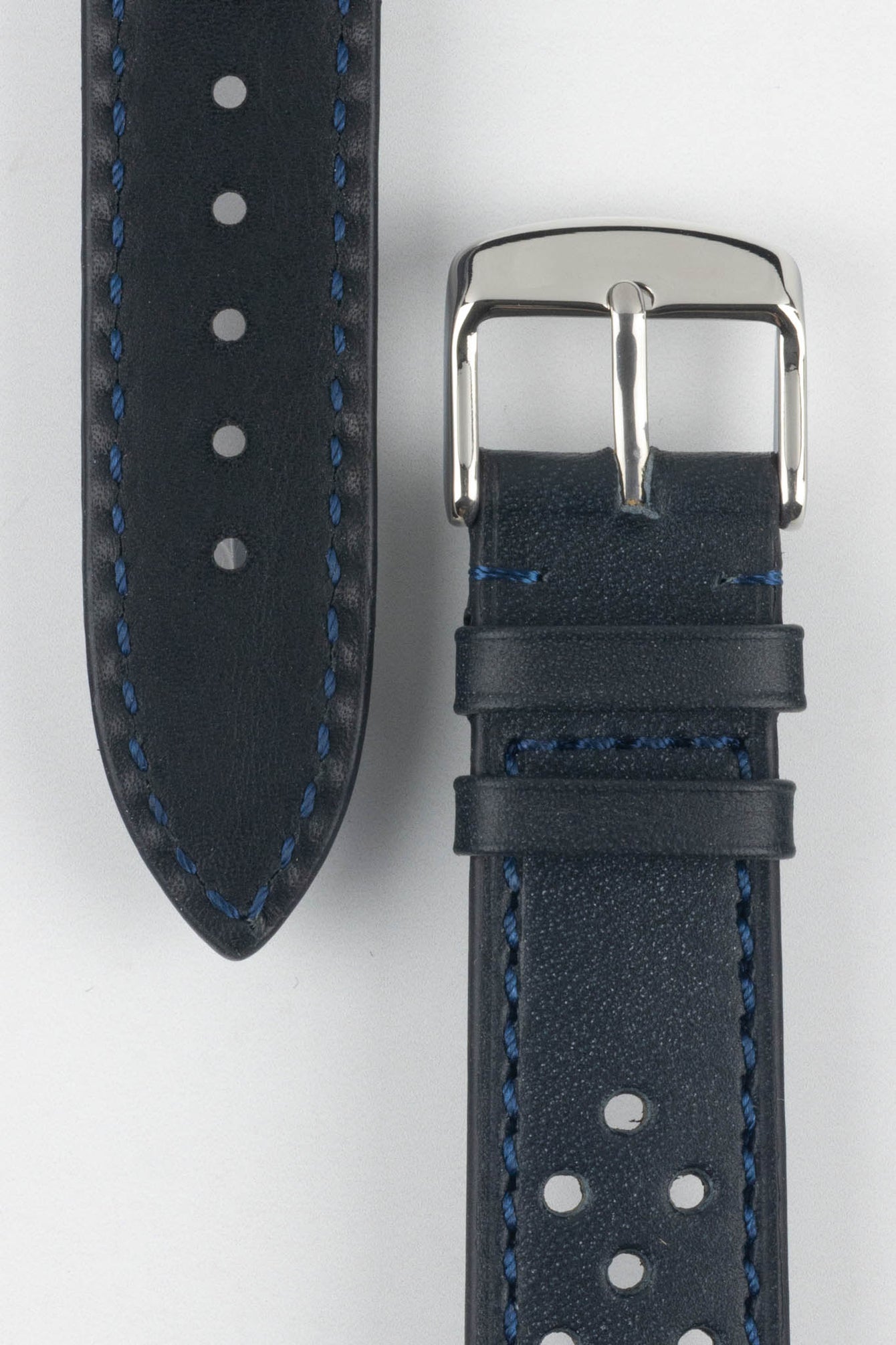 Close-up product shot of a blue-black perforated leather watch strap paired with a black underside strap segment, both featuring blue contrast stitching. A polished silver pin buckle is attached to the main strap, and the components are arranged on a plain light background to highlight the leather texture, perforations, stitching detail, and buckle hardware.
