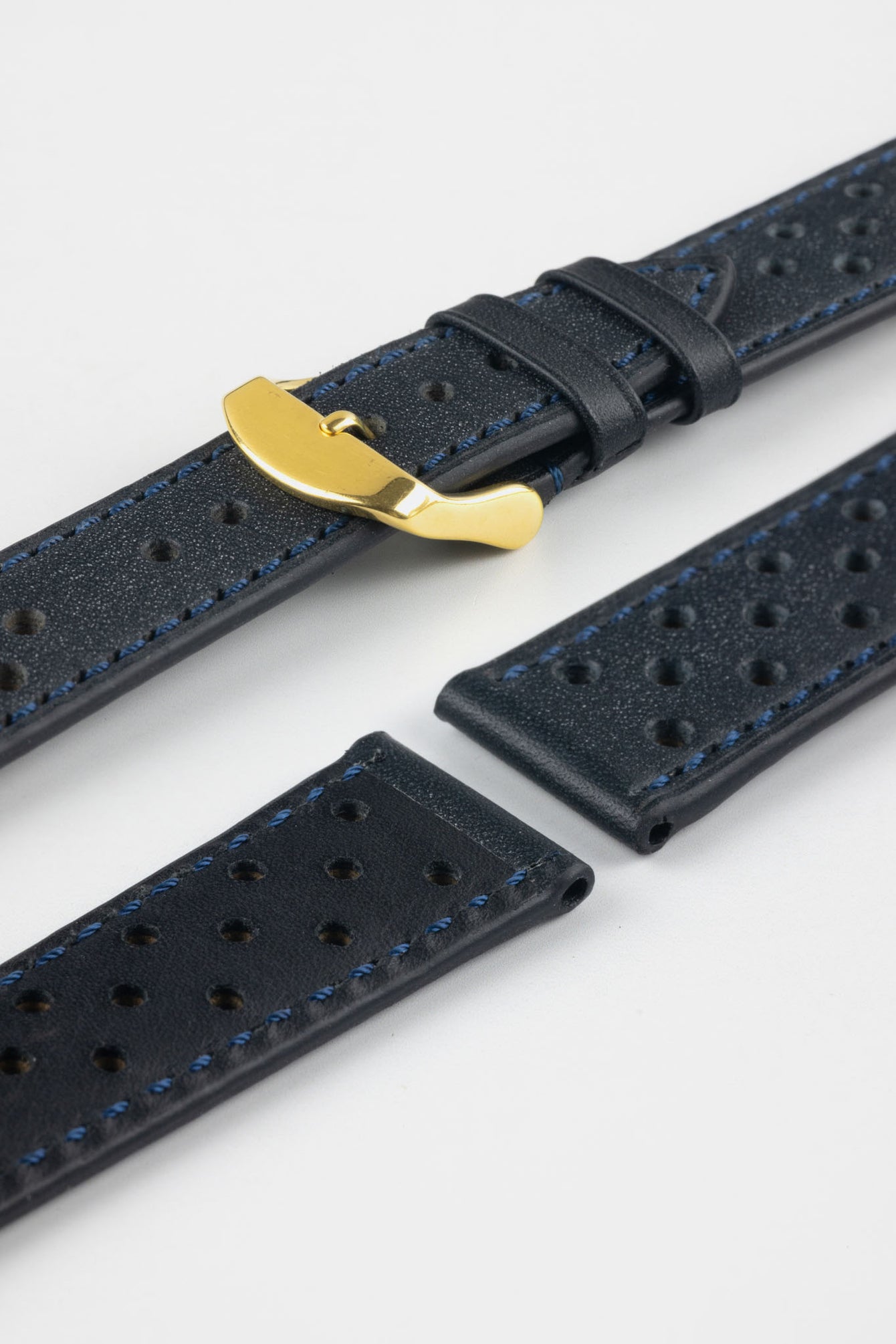 Close-up product shot of blue-black perforated leather watch strap components with blue contrast stitching, including one fitted with a polished gold-tone deployment clasp. Additional strap sections, including one with a black underside, are arranged on a plain light background, highlighting the leather texture, perforations, stitching detail, and gold clasp hardware.
