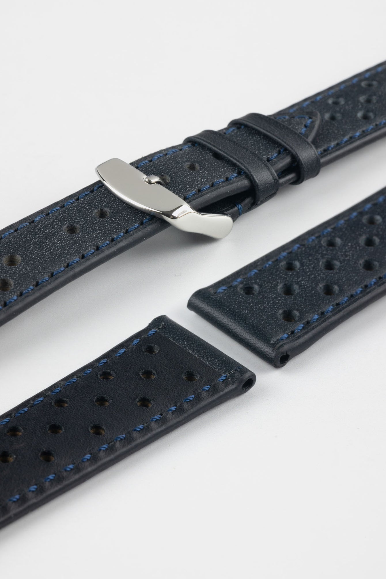 Close-up product shot of blue-black perforated leather watch strap components with blue contrast stitching, including one fitted with a polished silver deployment clasp. Additional strap sections, including one with a black underside, are arranged on a plain light background, highlighting the textured leather, perforations, stitching detail, and metal clasp hardware.