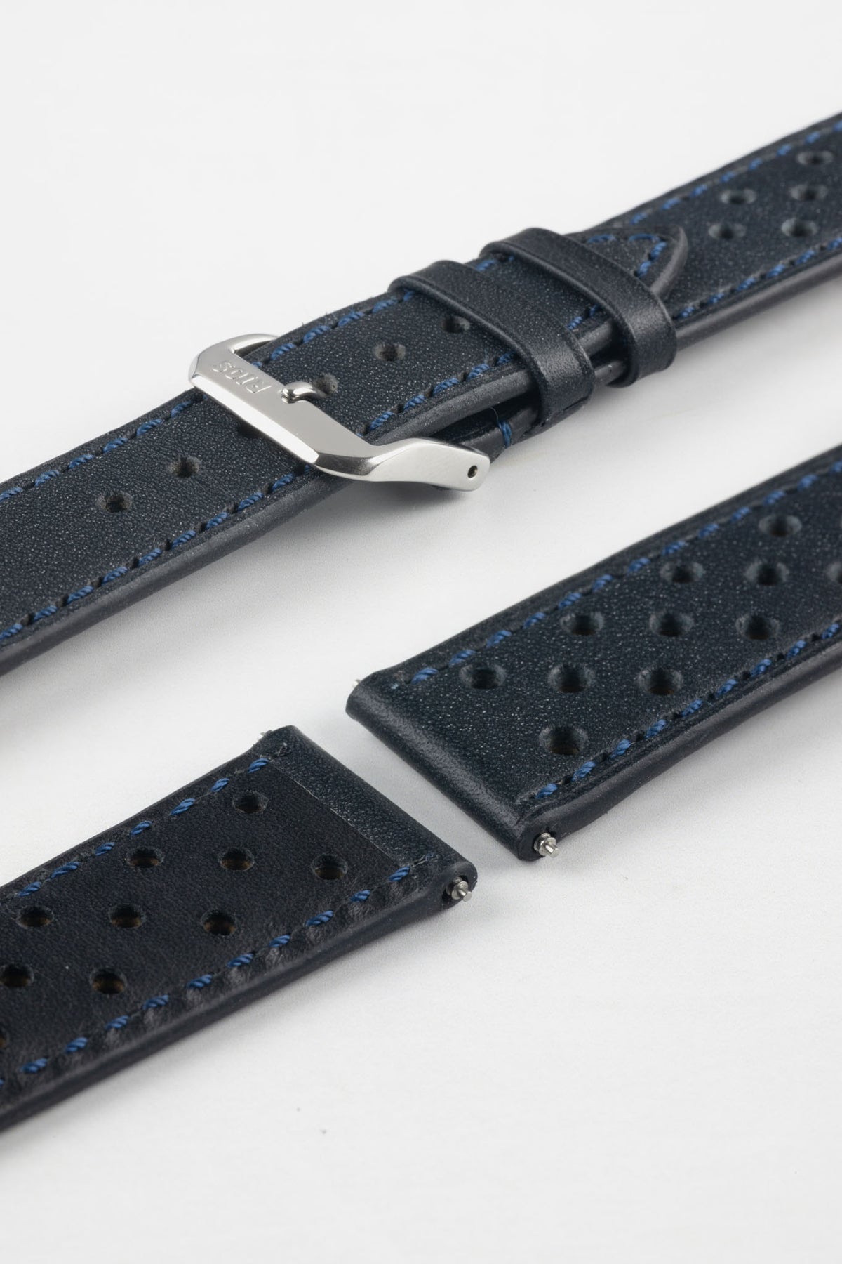 Close-up detail view of an ocean blue perforated leather rally watch strap displayed with one strap section detached, featuring matching blue stitching, multiple motorsport-inspired perforations, dual keeper loops, a polished stainless steel RIOS buckle, quick-release spring bars, and a black leather lining. Displayed on a light grey background to highlight the premium handcrafted construction, contrasting underside, and vintage racing-inspired design.