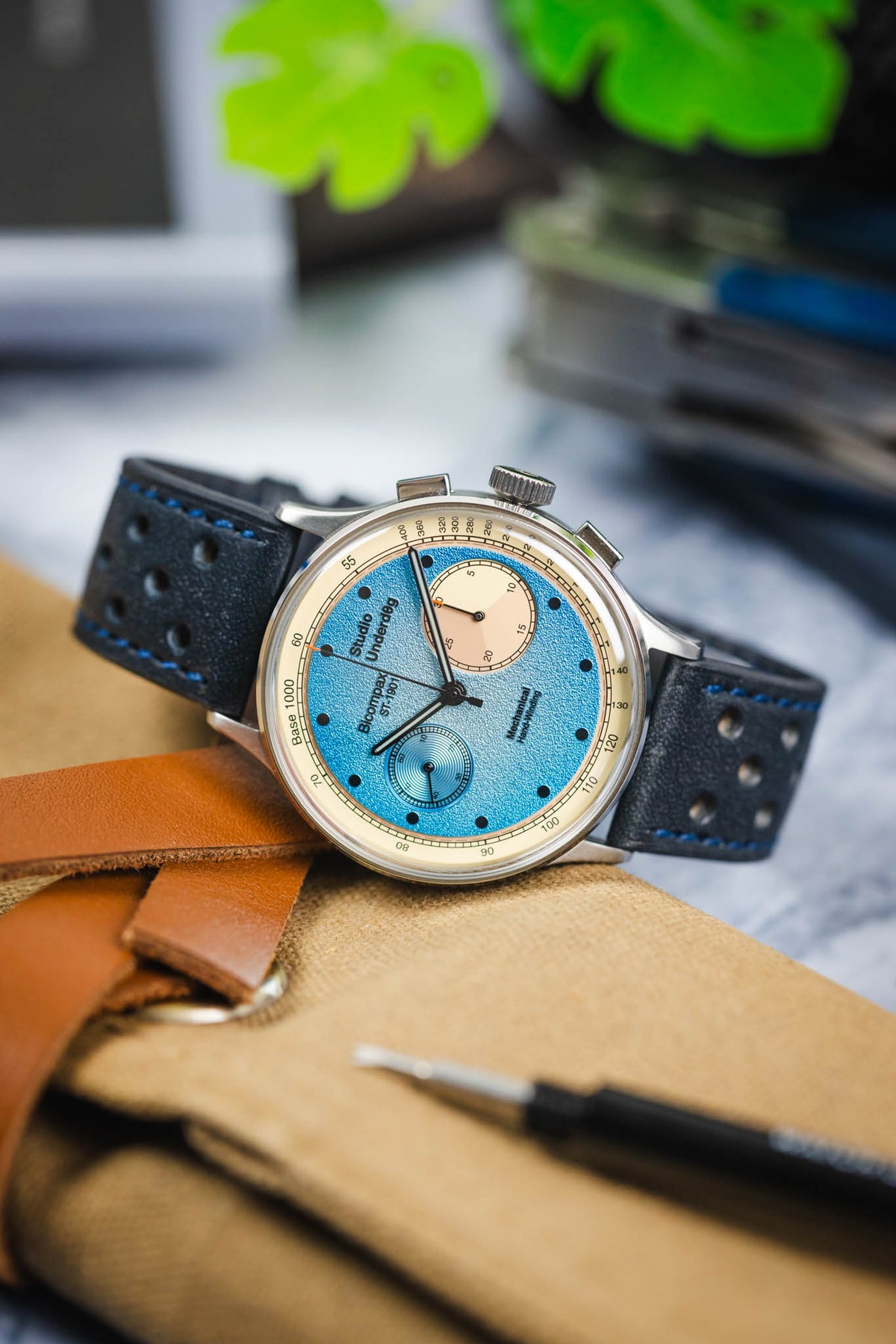 Close-up of a Studio Underd0g Bicompax mechanical chronograph watch featuring a textured light blue dial with contrasting cream sub-dials, polished stainless steel case, and an ocean blue perforated leather rally strap with matching blue stitching. Displayed on a canvas tool roll alongside leather accessories and a watch tool, the image highlights the strap's soft nubuck-style texture, motorsport-inspired perforations, and premium handcrafted construction.