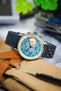 Close-up of a Studio Underd0g Bicompax mechanical chronograph watch featuring a textured light blue dial with contrasting cream sub-dials, polished stainless steel case, and an ocean blue perforated leather rally strap with matching blue stitching. Displayed on a canvas tool roll alongside leather accessories and a watch tool, the image highlights the strap's soft nubuck-style texture, motorsport-inspired perforations, and premium handcrafted construction.