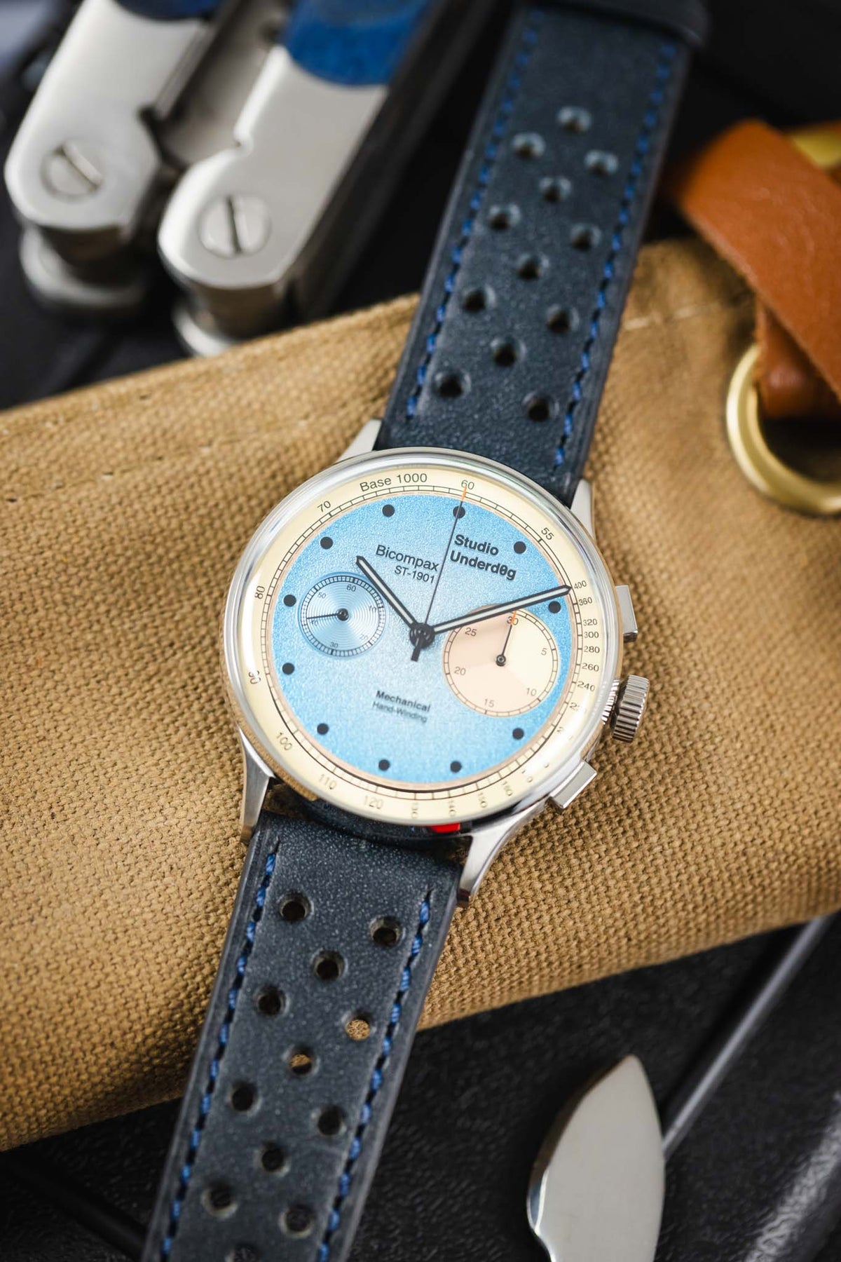 Close-up of a Studio Underd0g Bicompax mechanical chronograph watch featuring a textured light blue dial with contrasting cream sub-dials, polished stainless steel case, and an ocean blue perforated leather rally strap with matching blue stitching. Displayed on a canvas tool roll alongside a multitool and leather accessories, the image highlights the strap's soft nubuck-style texture, motorsport-inspired perforations, vibrant blue colour, and premium handcrafted construction.