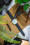 Flat lay of a mechanical chronograph wristwatch with a textured blue dial and cream outer ring, paired with a dark perforated leather strap with blue stitching, positioned diagonally on a tan canvas watch roll. Surrounding items include a spring bar tool labeled “WATCH OBSESSION,” a brown leather strap, sunglasses, and green plant leaves partially framing the scene, all set on a marble surface.