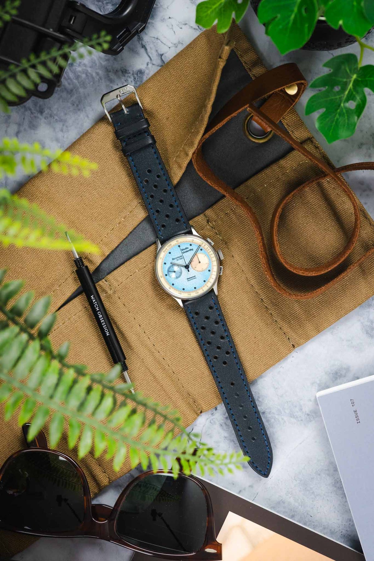 Vintage-style Studio Underd0g Bicompax chronograph watch with a light blue dial and cream sub-dials, fitted with an ocean blue perforated leather rally strap featuring matching blue stitching and a tapered profile. Displayed on a canvas tool roll alongside a Watch Obsession spring bar tool, sunglasses, leather accessories, and greenery, highlighting the strap's motorsport-inspired design, vibrant colour, and premium handcrafted construction.