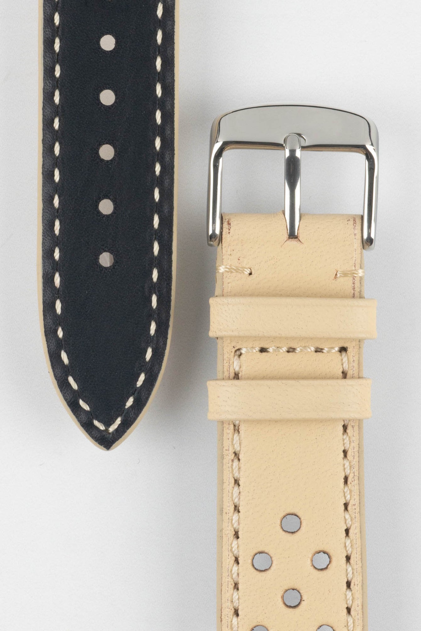 Close-up product shot of a beige perforated leather watch strap paired with a black underside strap segment, both featuring contrasting stitching. A polished silver pin buckle is attached to the beige strap, and the pieces are arranged on a plain light background to highlight texture, perforations, and finishing details.