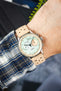Close-up of a wrist wearing a mechanical chronograph watch with a pale green textured dial and cream outer ring, featuring two subdials and black hands. The watch is paired with a beige perforated leather strap and worn over a blue-and-white plaid shirt cuff, with a softly blurred background.