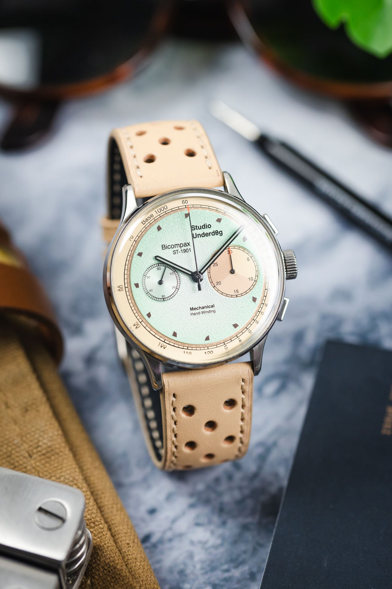 Close-up of a mechanical chronograph wristwatch with a pale green textured dial and cream outer ring, featuring two subdials and black hands. The watch is paired with a beige perforated leather strap and rests on a marble surface, with a tan canvas pouch, sunglasses, and a spring bar tool softly blurred in the background.