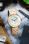 Close-up of a mechanical chronograph wristwatch with a pale green textured dial and cream outer ring, featuring two subdials and black hands. The watch is paired with a beige perforated leather strap and rests on a marble surface, with a tan canvas pouch, sunglasses, and a spring bar tool softly blurred in the background.