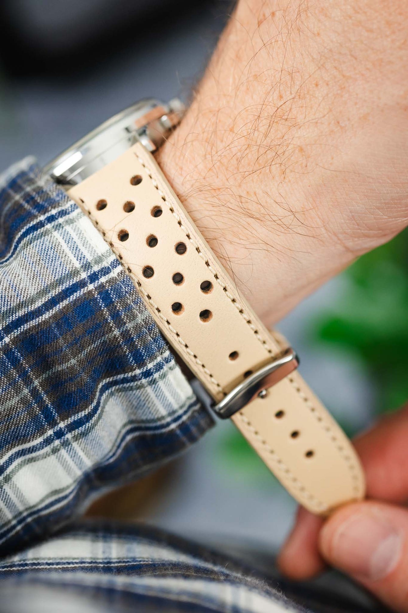 Close-up of a beige perforated leather watch strap worn on a wrist, showing detailed stitching and a silver buckle. The strap is paired with a watch partially visible at the edge of the frame and worn over a blue-and-white plaid shirt cuff, with a softly blurred background of greenery.