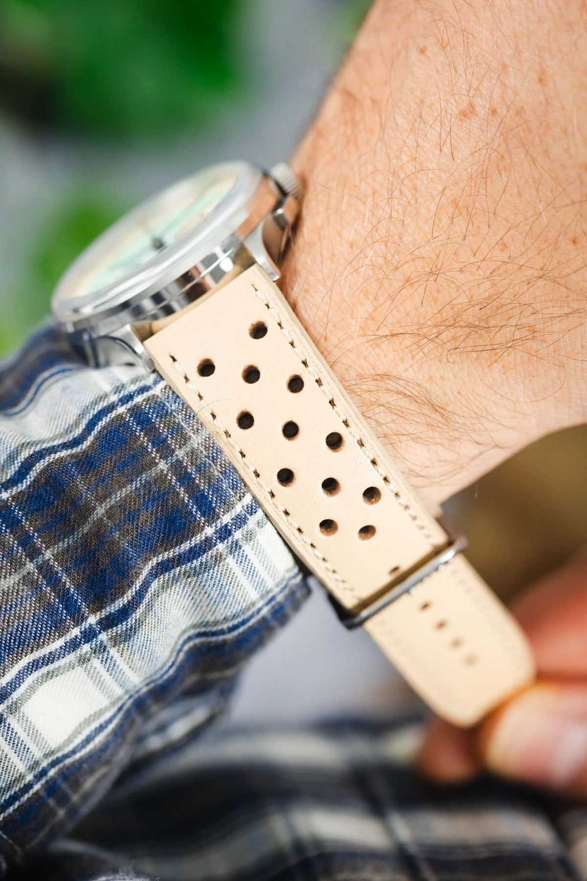 Close-up of a sand-coloured perforated leather rally watch strap worn on the wrist, featuring multiple racing-style perforations, cream contrast stitching, and a polished stainless steel buckle, paired with a blue plaid shirt cuff to highlight the strap's vintage motorsport-inspired design and premium handcrafted leather construction.