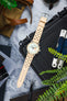 Flat lay of a mechanical chronograph wristwatch with a light dial and two subdials, paired with a beige perforated leather strap, positioned diagonally across a black case on a marble surface. Surrounding items include a tan canvas watch roll with brown leather straps, a multi-tool, a spring bar tool partially visible, sunglasses, and green plant leaves framing the scene.