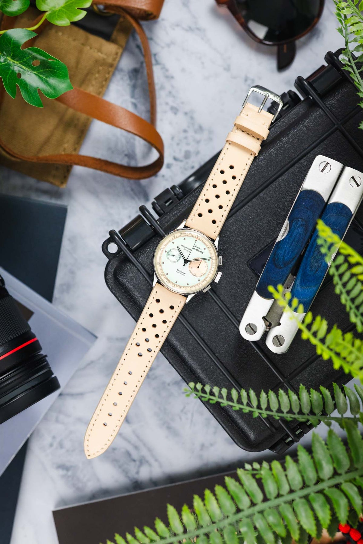 Vintage-style Studio Underd0g chronograph watch with a pastel green and cream dial, fitted with a sand-coloured perforated leather rally strap, displayed on a black equipment case alongside a multitool, camera lens, sunglasses, leather accessories, and greenery, highlighting the strap's motorsport-inspired design and premium handcrafted leather construction.