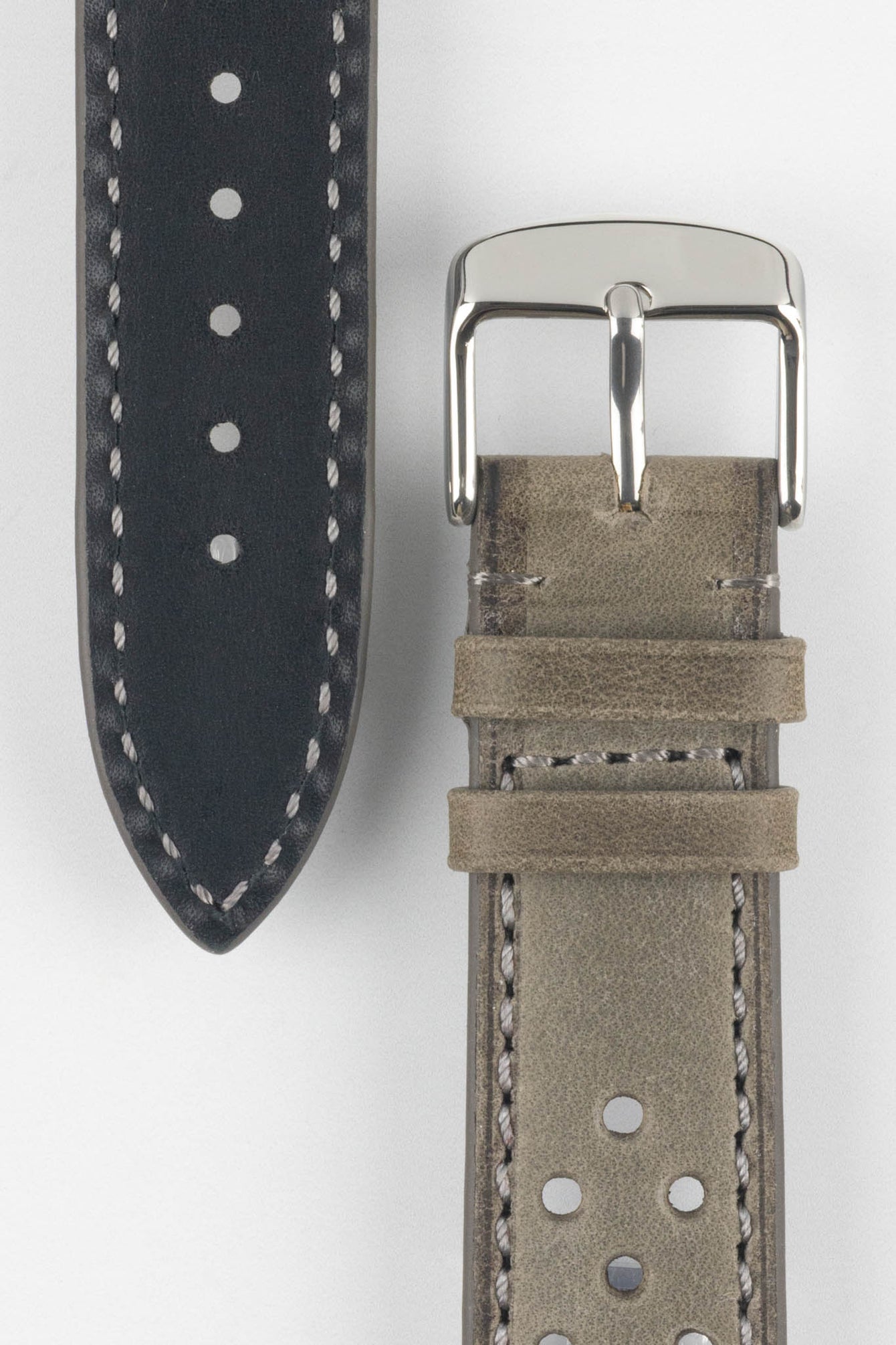 Close-up product shot of a grey suede perforated leather watch strap alongside a black underside strap segment, both with contrasting stitching. A polished silver pin buckle is attached to the grey strap, and the components are arranged on a plain light background to highlight texture, perforations, and finishing details.