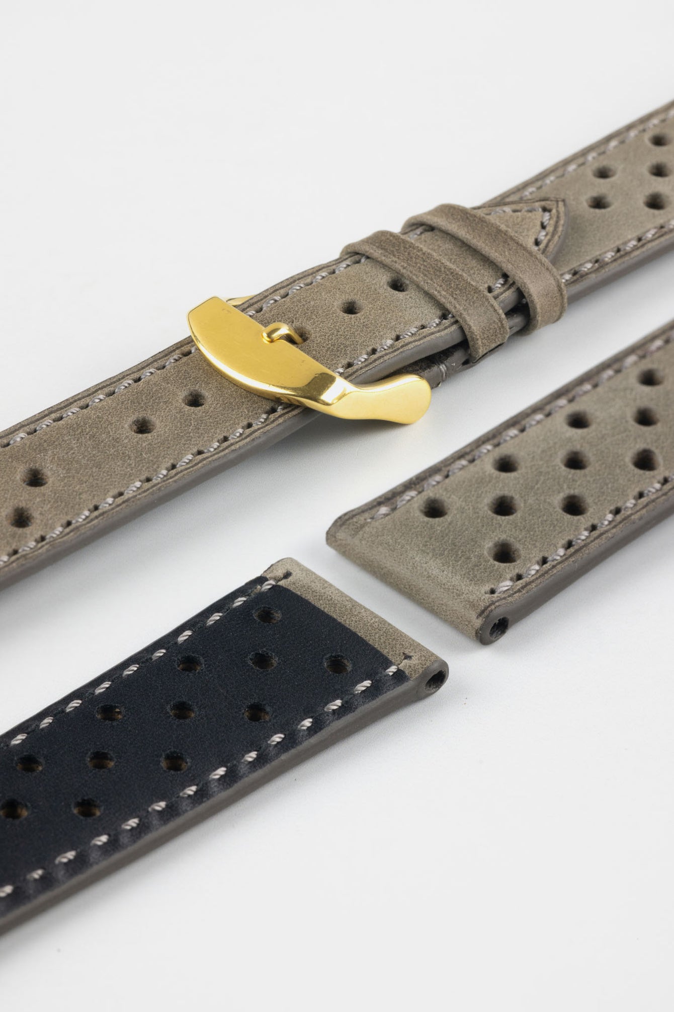 Close-up product shot of grey suede perforated leather watch strap components with light contrast stitching, including one fitted with a polished gold-tone deployment clasp. Additional strap pieces, including one with a black underside, are arranged on a plain light background to highlight the perforations, soft texture, and metal hardware.