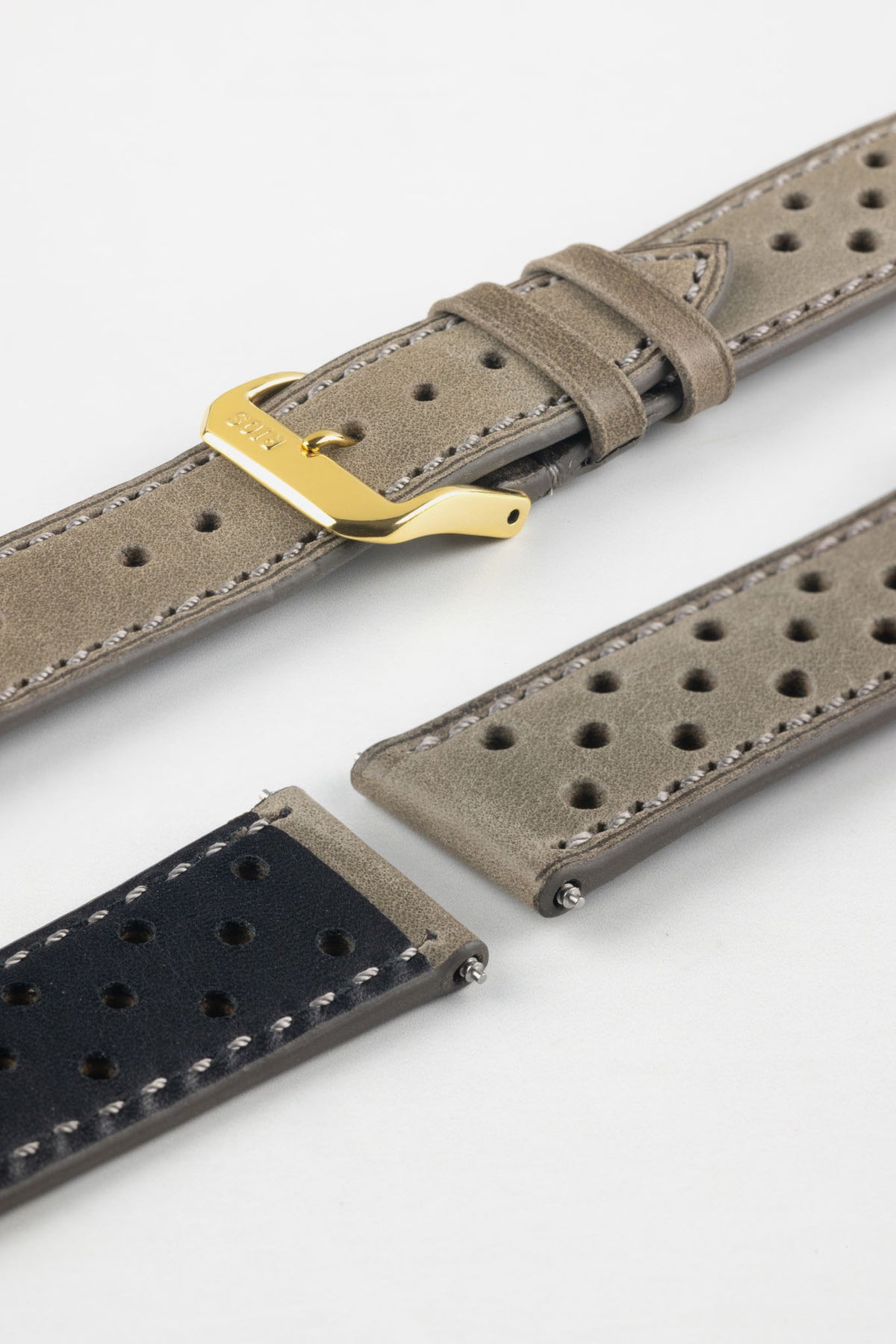 Close-up detail view of a stone grey perforated leather rally watch strap displayed with one strap section detached, featuring a soft nubuck-style finish, tonal grey stitching, multiple motorsport-inspired perforations, dual keeper loops, a polished gold-tone RIOS buckle, quick-release spring bars, and a black leather lining. Displayed on a light grey background to highlight the premium handcrafted construction, contrasting underside, and vintage racing-inspired design.