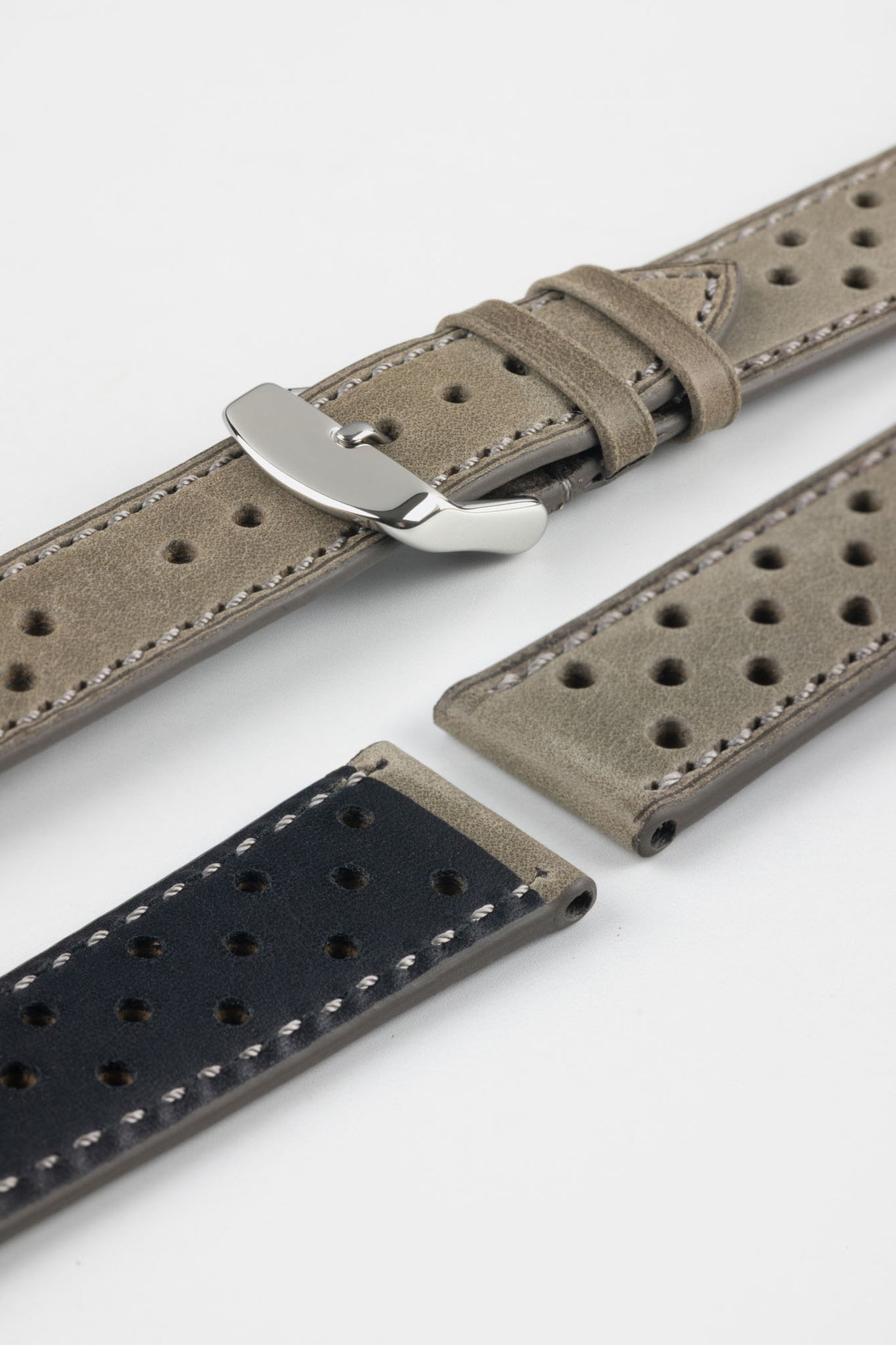 Close-up product shot of grey suede perforated leather watch strap components with light contrast stitching, including one fitted with a polished silver deployment clasp. Additional strap sections, including one with a black underside, are arranged on a plain light background to highlight the soft texture, perforations, and metal hardware details.