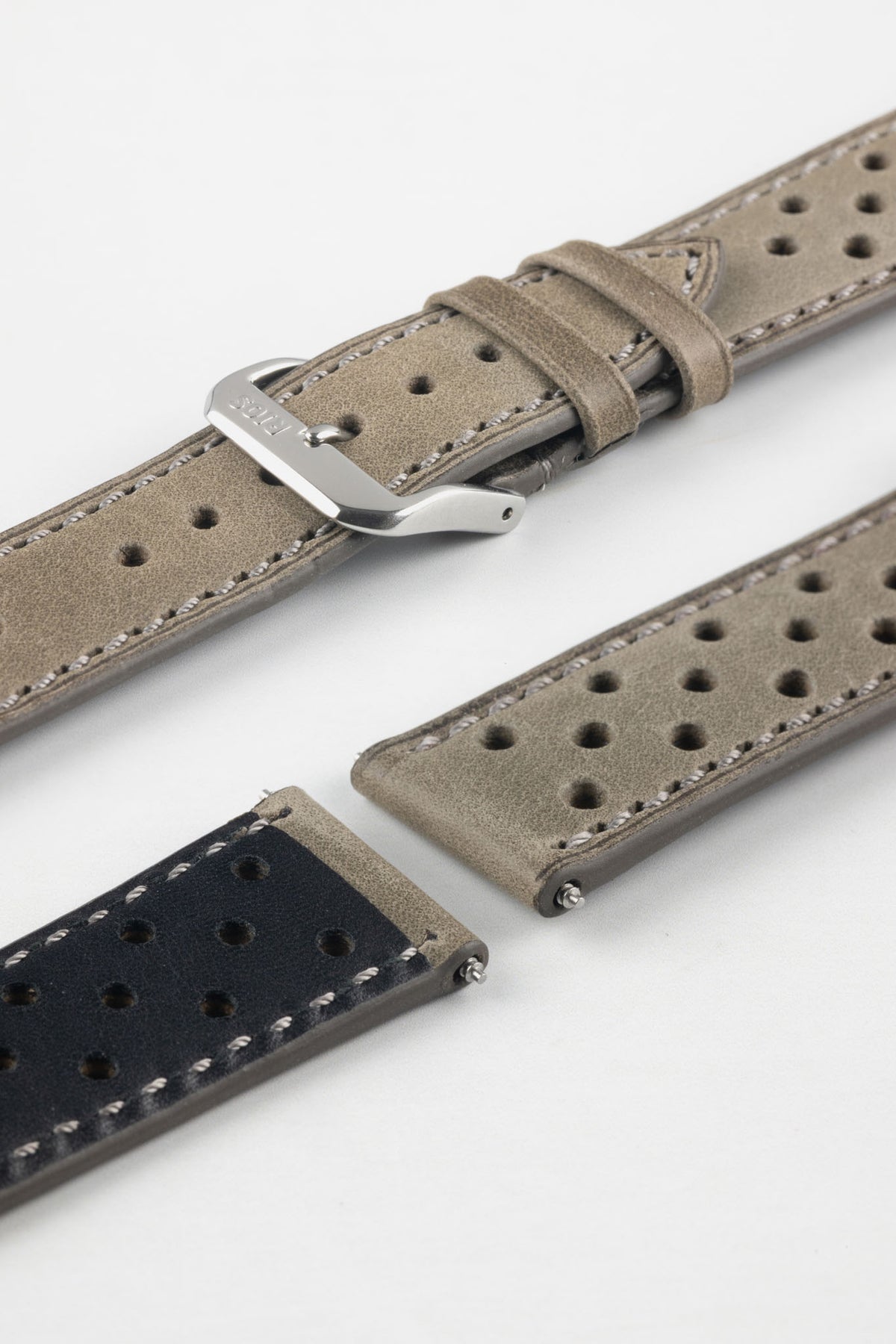 Close-up detail view of a stone grey perforated leather rally watch strap displayed with one strap section detached, featuring a soft nubuck-style finish, tonal grey stitching, multiple motorsport-inspired perforations, dual keeper loops, a polished stainless steel RIOS buckle, quick-release spring bars, and a black leather lining. Displayed on a light grey background to highlight the premium handcrafted construction, contrasting underside, and vintage racing-inspired design.
