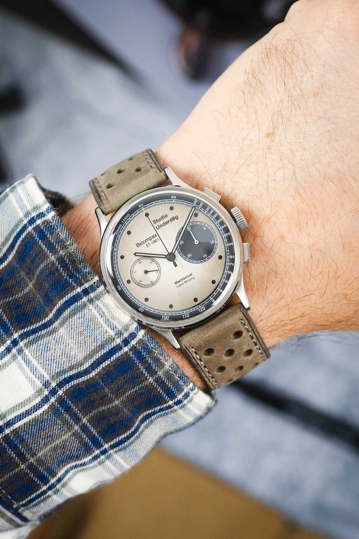 Studio Underd0g Bicompax mechanical chronograph watch worn on the wrist, featuring a textured cream dial with contrasting dark grey sub-dials, polished stainless steel case, and a stone grey perforated leather rally strap with tonal stitching. Paired with a blue plaid shirt cuff, the image highlights the strap's soft nubuck-style texture, motorsport-inspired perforations, and premium handcrafted construction.