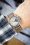 Studio Underd0g Bicompax mechanical chronograph watch worn on the wrist, featuring a textured cream dial with contrasting dark grey sub-dials, polished stainless steel case, and a stone grey perforated leather rally strap with tonal stitching. Paired with a blue plaid shirt cuff, the image highlights the strap's soft nubuck-style texture, motorsport-inspired perforations, and premium handcrafted construction.