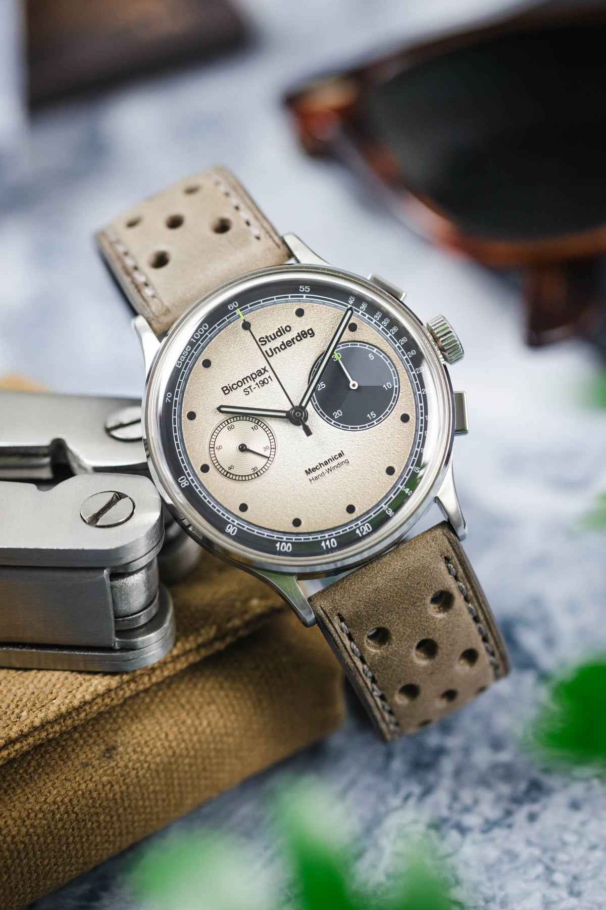 Close-up of a Studio Underd0g Bicompax mechanical chronograph watch featuring a textured cream dial with contrasting dark grey sub-dials, polished stainless steel case, and a stone grey perforated leather rally strap with tonal stitching. Displayed alongside a multitool, sunglasses, and canvas accessories, the image highlights the strap's soft nubuck-style texture, motorsport-inspired perforations, and premium handcrafted construction.
