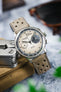 Close-up of a Studio Underd0g Bicompax mechanical chronograph watch featuring a textured cream dial with contrasting dark grey sub-dials, polished stainless steel case, and a stone grey perforated leather rally strap with tonal stitching. Displayed alongside a multitool, sunglasses, and canvas accessories, the image highlights the strap's soft nubuck-style texture, motorsport-inspired perforations, and premium handcrafted construction.