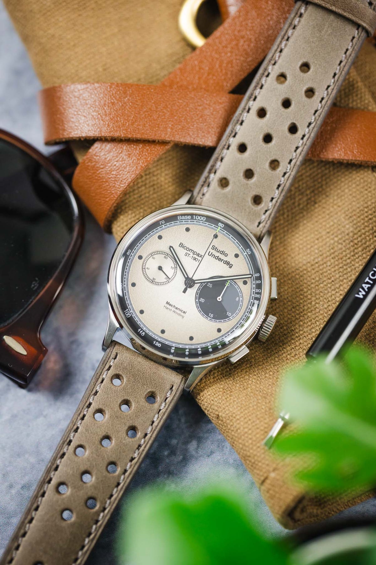 Close-up of a Studio Underd0g Bicompax mechanical chronograph watch featuring a textured cream dial with contrasting dark grey sub-dials, polished stainless steel case, and a stone grey perforated leather rally strap with tonal stitching. Displayed atop a canvas pouch alongside sunglasses, a leather strap, and a watch tool, the image highlights the strap's soft nubuck-like texture, motorsport-inspired perforations, and premium handcrafted construction.