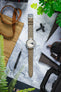 Flat lay of a mechanical chronograph watch with a beige dial and perforated grey leather strap placed vertically on a marble surface, surrounded by watch tools, sunglasses, a tan canvas roll with brown leather straps, and green plant leaves partially framing the scene.