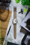 Vintage-style Studio Underd0g Bicompax chronograph watch with a cream dial and contrasting dark grey sub-dial, fitted with a stone grey perforated leather rally strap featuring tonal stitching and a tapered profile. Displayed on a marble-effect surface alongside a spring bar tool, sunglasses, camera equipment, canvas accessories, and greenery, highlighting the strap's motorsport-inspired design, soft suede-like texture, and premium handcrafted construction.