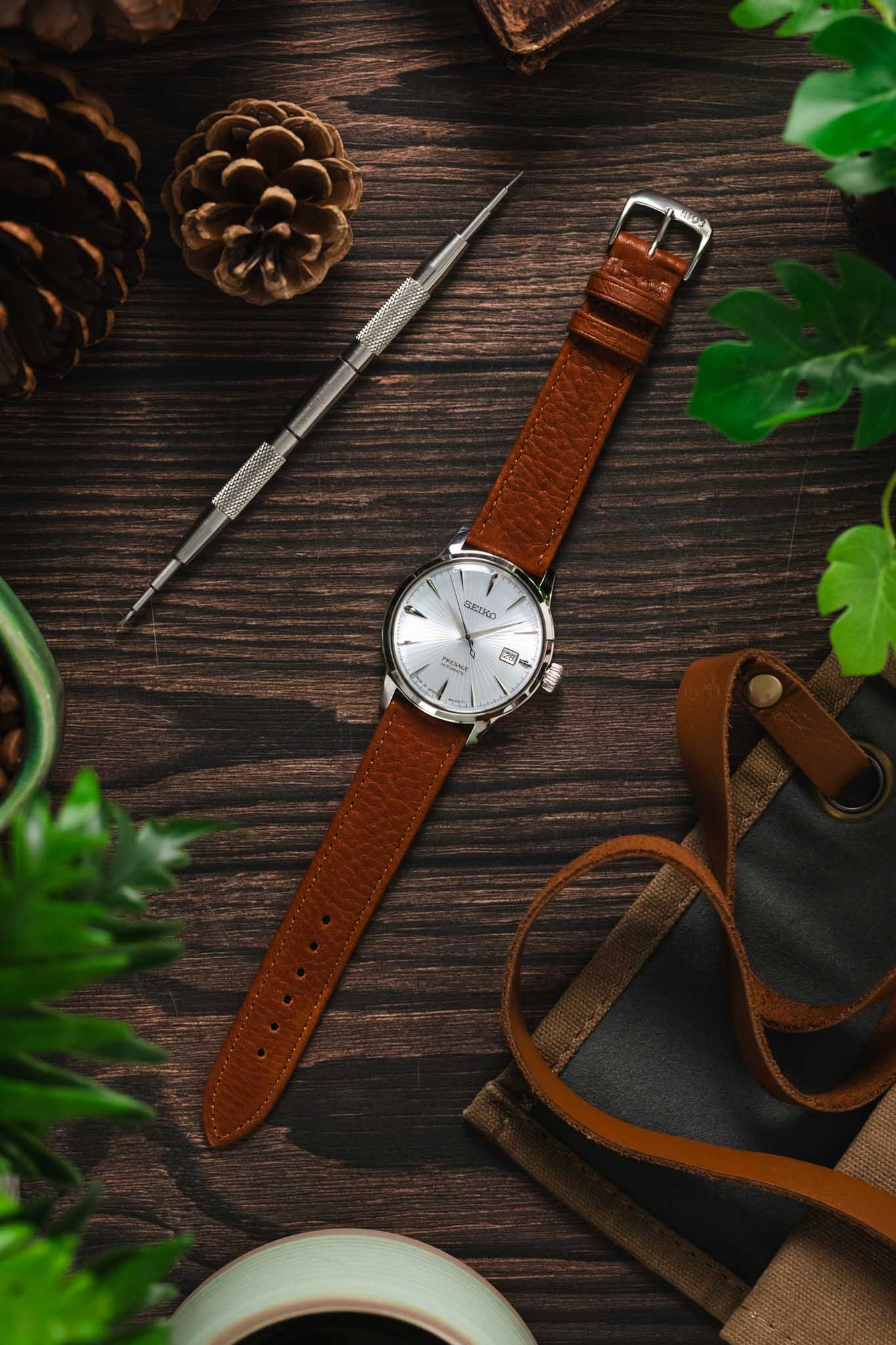 Moody lay flat scene with Seiko cocktail time watch on cognac Rios 1931 waging strap on dark wood with pine cones and plants