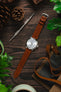 Moody lay flat scene with Seiko cocktail time watch on cognac Rios 1931 waging strap on dark wood with pine cones and plants