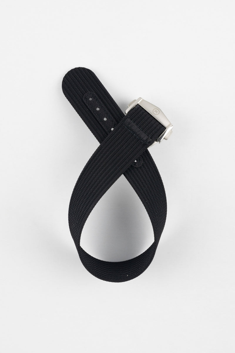 A top-down shot of a black ribbed-nylon watch strap coiled into a loop against a white background, featuring a brushed stainless steel buckle with a circular logo.
