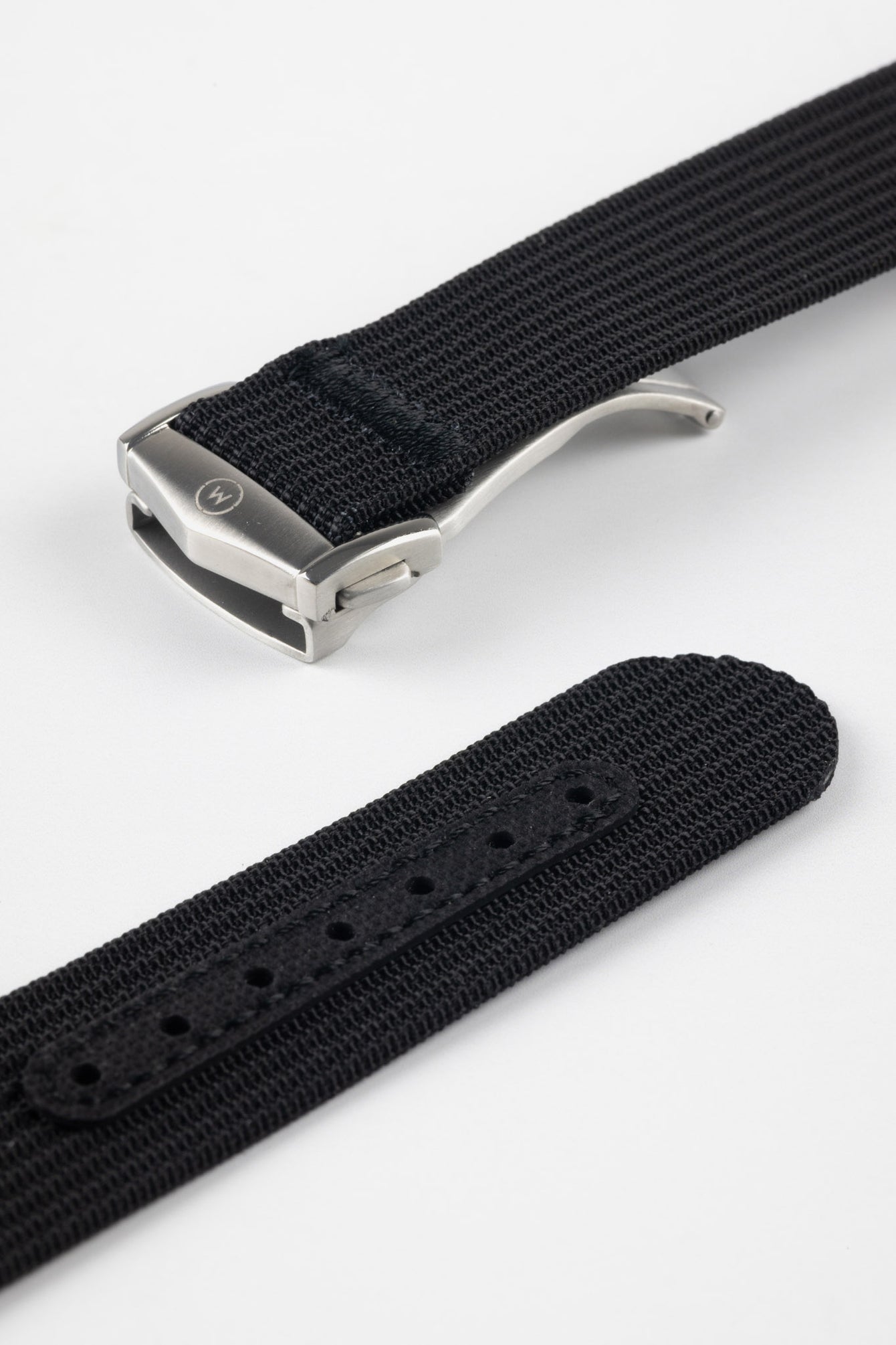 A close-up, angled shot of a black ribbed-nylon watch strap showing both the brushed stainless steel deployment buckle and the adjustment holes. The image highlights the fine woven texture of the fabric and the precision stitching near the metal clasp.