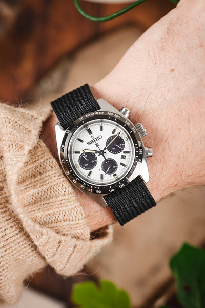 A close-up shot of a person's wrist wearing a Seiko Speedtimer watch with a white "panda" dial. The watch is paired with a black ribbed-nylon strap and is worn over the sleeve of a beige cable-knit sweater, creating a cozy, outdoor-inspired look.