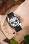 A close-up shot of a person's wrist wearing a Seiko Speedtimer watch with a white "panda" dial. The watch is paired with a black ribbed-nylon strap and is worn over the sleeve of a beige cable-knit sweater, creating a cozy, outdoor-inspired look.