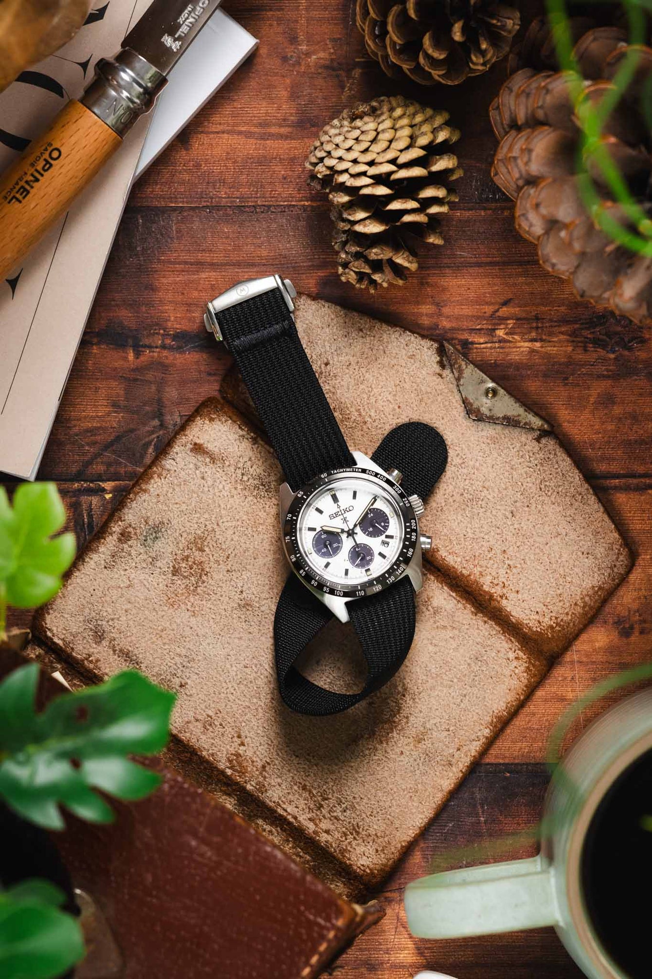 A top-down lifestyle shot of a Seiko Prospex Speedtimer chronograph watch with a white dial and black sub-dials, fitted on a black ribbed-nylon strap. The watch rests on a rustic leather pouch atop a wooden surface, surrounded by pine cones, an Opinel folding knife, and green foliage.