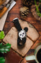 A top-down lifestyle shot of a Seiko Prospex Speedtimer chronograph watch with a white dial and black sub-dials, fitted on a black ribbed-nylon strap. The watch rests on a rustic leather pouch atop a wooden surface, surrounded by pine cones, an Opinel folding knife, and green foliage.