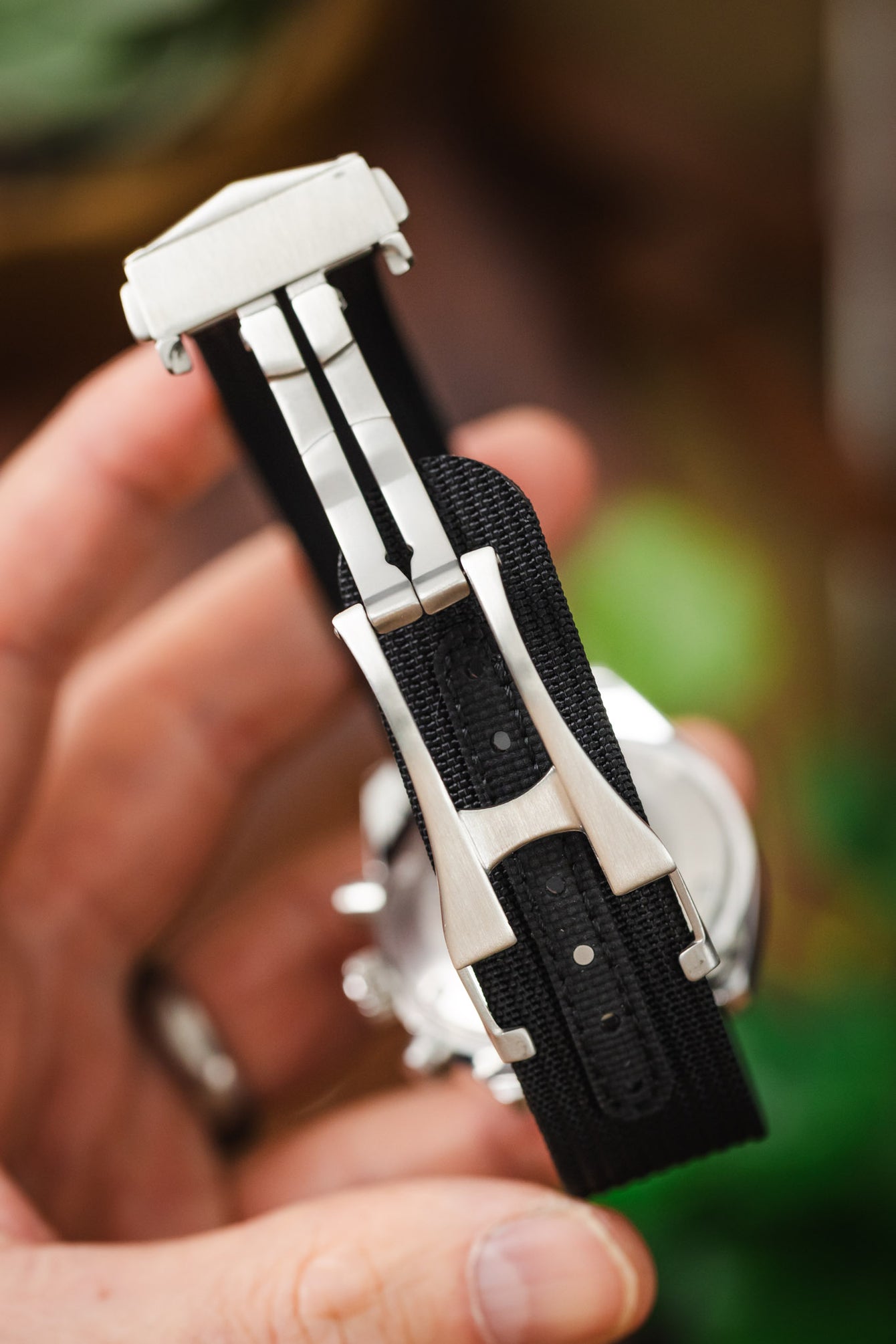 Close-up of a black textured fabric watch strap fitted with a brushed stainless steel deployant clasp, held in a hand, with the clasp mechanism and adjustment holes clearly visible against a softly blurred wooden background with hints of green foliage.