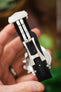 Close-up of a black textured fabric watch strap fitted with a brushed stainless steel deployant clasp, held in a hand, with the clasp mechanism and adjustment holes clearly visible against a softly blurred wooden background with hints of green foliage.